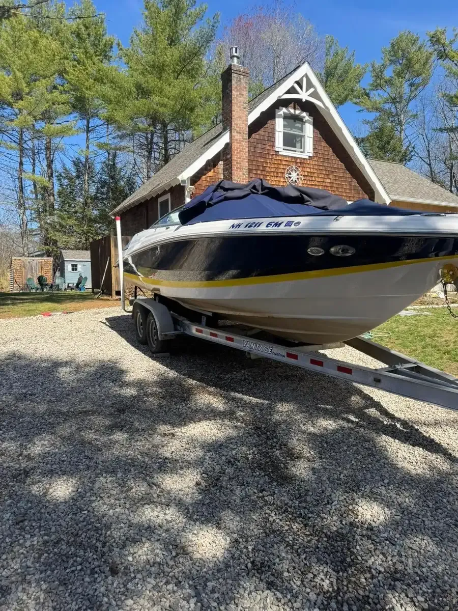 A navy bowrider under its cover on a galvanised trailer in a gravel drive outside a shingled New Hampshire house, trees still bare in early spring.