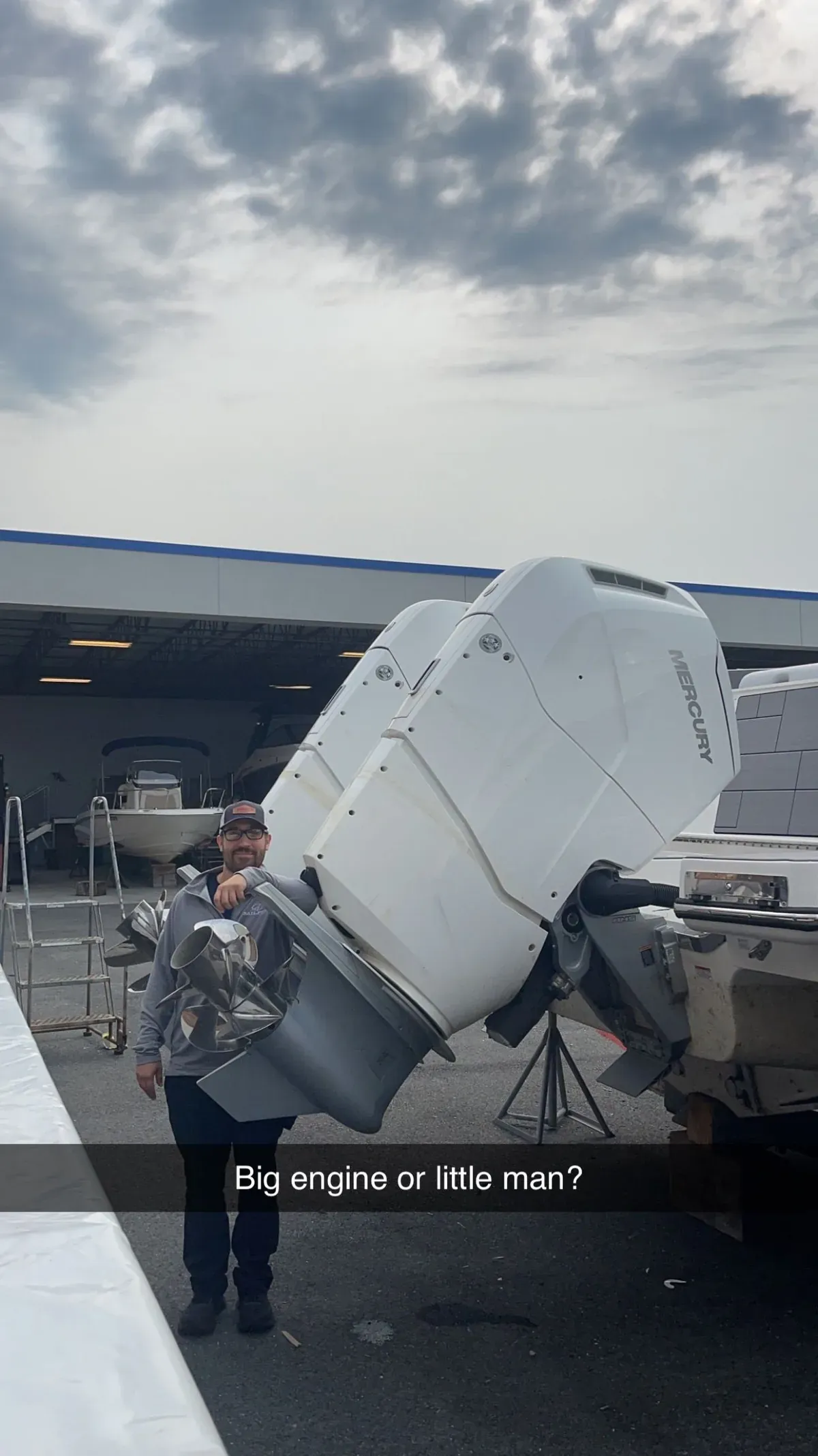A technician carrying a Mercury outboard lower unit and stainless propeller past a boat with twin white Mercury outboards tilted up.