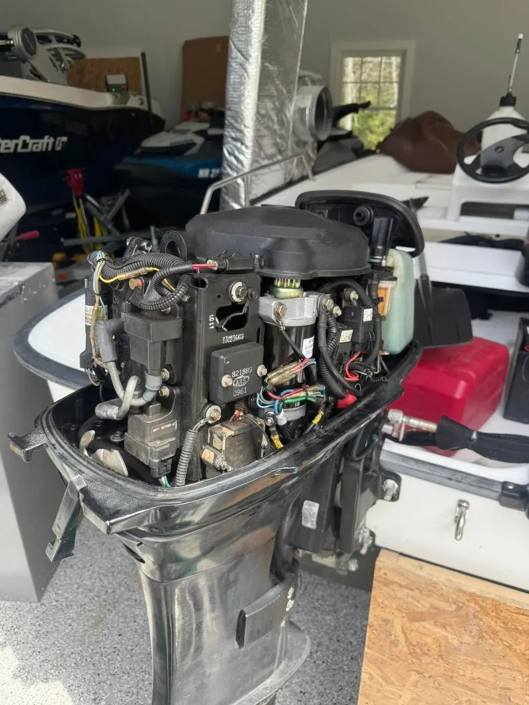 An outboard with its cowling removed, showing the powerhead, ignition coils, wiring harness and fuel pump, on the bench in a garage.